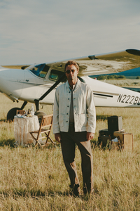 Man wearing a utility-style jacket, collared shirt, and relaxed trousers, standing outdoors in front of a vintage truck with mountain scenery in the background.