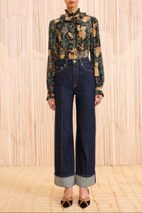 Model wearing a floral blouse and dark blue jeans against a wooden paneled wall.