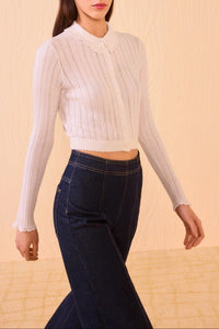 Woman wearing a white ribbed long-sleeve top and dark blue jeans against a beige background