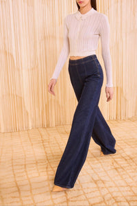 Woman wearing a white long-sleeve top and dark blue jeans against a wooden paneled wall.